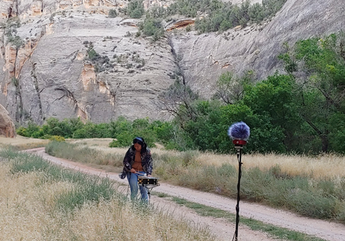 Image: Susie Ibarra (AWAW EAG '23) recording a snare in the echos of a whispering cave canyon at Dinosaur Monument Park, Colorado and Utah border, Photo Credit: Tessa Fuqua