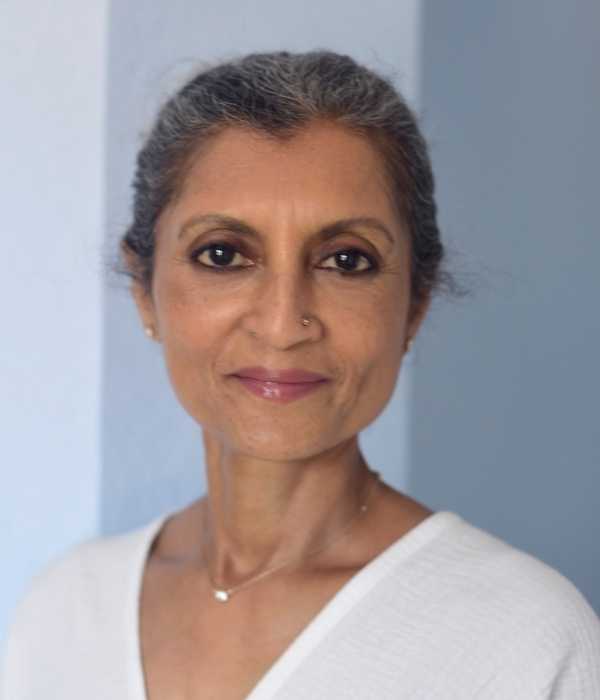 Smiling person with grey hair pulled back, tan skin and white top.