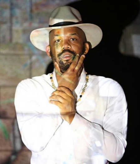 Bean, Outspoken Headshot Person with warm skin and facial hair, wearing hat, and holding their face with a projection overlay.