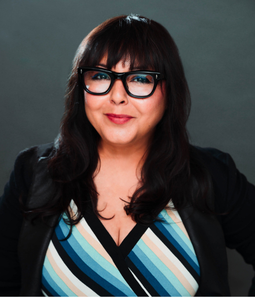 Smiling person with dark, long hair and glasses in front of a dark background.