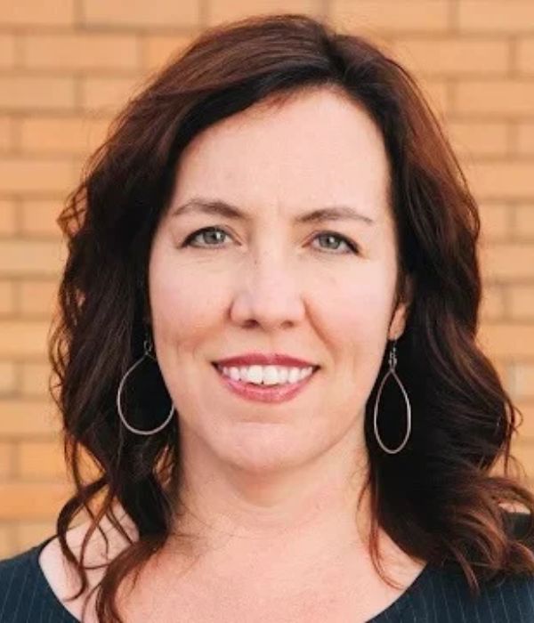 Smiling person with long, auburn hair against a brick background.