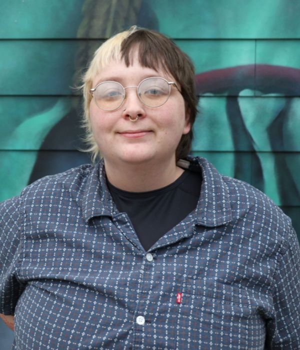 Smiling person with blonde and auburn short hair, glasses, and wearing a check shirt standing in front of a painted wall.