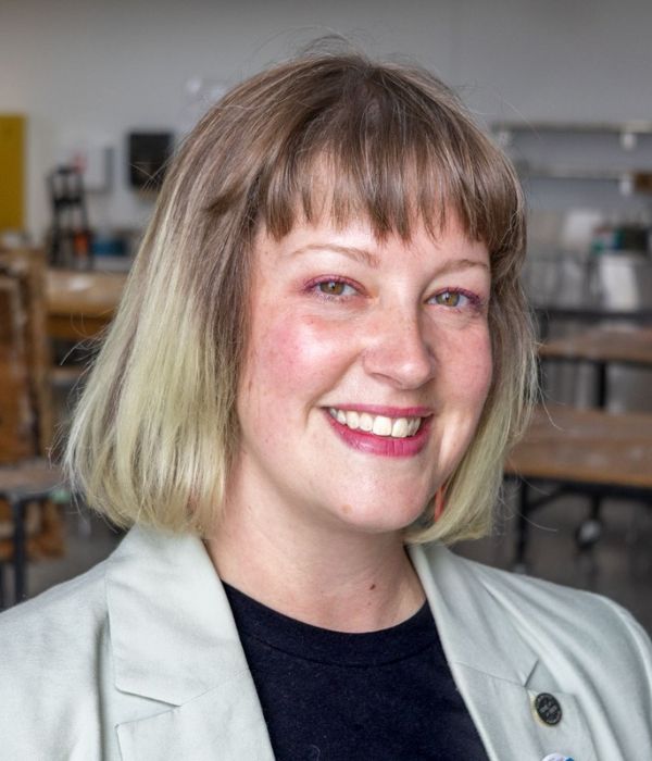 Smiling person with short, blonde hair with an art studio in the background