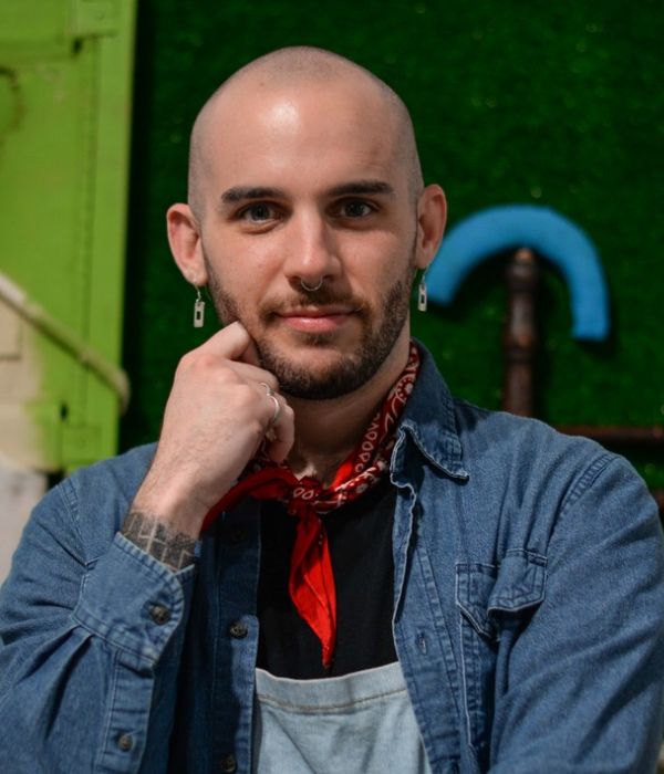 Smiling person with shaved head and facial hair, wearing a denim shirt and earrings.